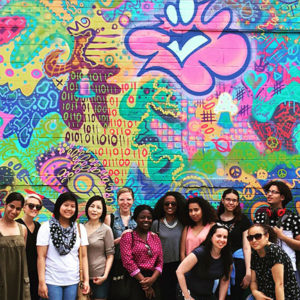 Group of high school students with their mentors smiling in front of large graffiti mural